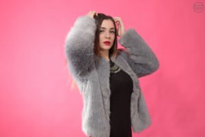 Portréfotó fiatal nőről rózsaszín háttér előtt, szürke szőrmekabátban és elegáns fekete ruhában. Stílusos portréfotózás divatfotós megközelítéssel.