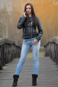 Kültéri fotózás fiatal nővel, fekete bőrdzsekiben, világoskék farmerben és bakancsban. Háttere egy fa híd és természetes környezet, meleg tónusú naplementei fényekkel. Ideális portré és divatfotózáshoz.