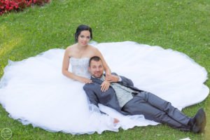 Esküvői pár a természetben, a menyasszony hófehér ruhájának szoknyája körülöleli a zöld füvet, miközben a vőlegény elegáns szürke öltönyben pihen mellette. Romantikus esküvői fotózás, természetes fényekkel, tökéletes pillanat megörökítése esküvői albumhoz.