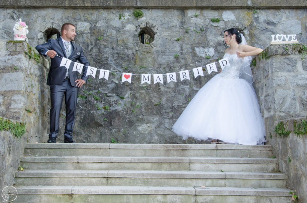 Friss házasok vicces pillanata esküvői fotózáson, kőfal előtt. A pár egy "Just Married" feliratú zászlót tart a kezében, mosolyogva és boldogan. A menyasszony elegáns fehér ruhában, a vőlegény stílusos öltönyben.