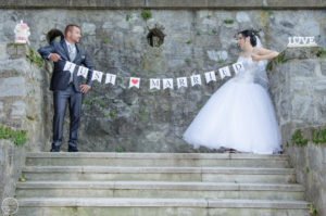Friss házasok vicces pillanata esküvői fotózáson, kőfal előtt. A pár egy "Just Married" feliratú zászlót tart a kezében, mosolyogva és boldogan. A menyasszony elegáns fehér ruhában, a vőlegény stílusos öltönyben.
