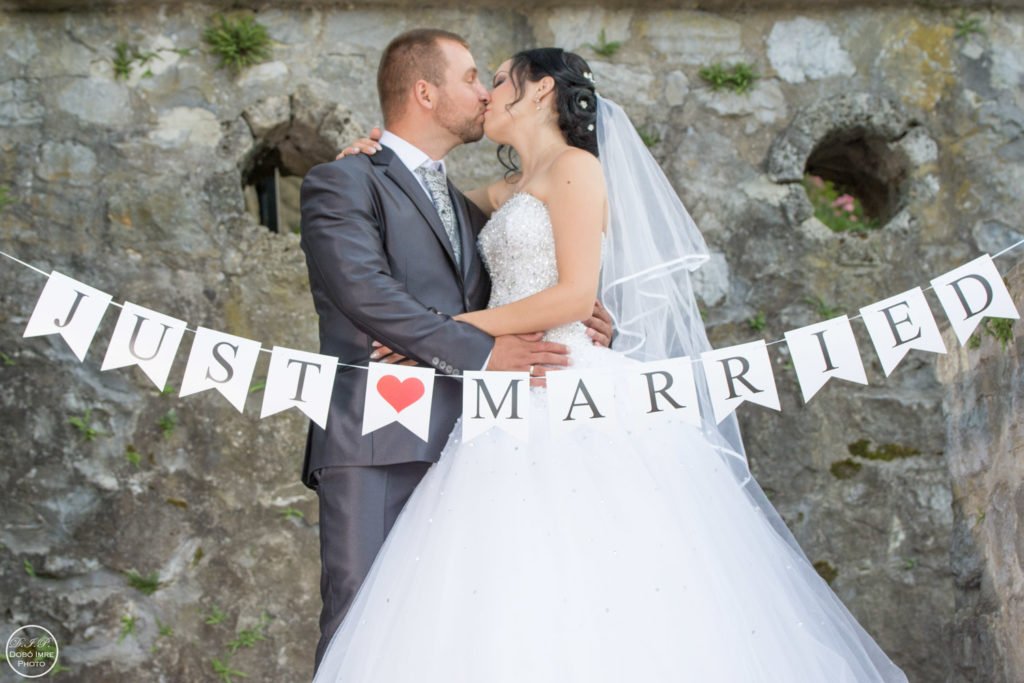 Esküvői fotózás - Debrecen. Friss házas pár csókja esküvői fotózáson, háttérben rusztikus kőfal. A pár előtt „Just Married” feliratú dekoráció, a menyasszony elegáns fehér ruhában és fátyolban, a vőlegény klasszikus szürke öltönyben. Tökéletes esküvői emlékkép.