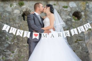 Esküvői fotózás - Debrecen. Friss házas pár csókja esküvői fotózáson, háttérben rusztikus kőfal. A pár előtt „Just Married” feliratú dekoráció, a menyasszony elegáns fehér ruhában és fátyolban, a vőlegény klasszikus szürke öltönyben. Tökéletes esküvői emlékkép.