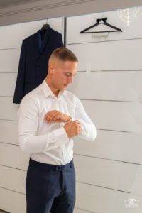 Vőlegény készülődik az esküvőre, fehér inget gombol fel, mellette elegáns kék öltöny a fogason. Esküvői fotózás részletfotó a vőlegény öltözködéséről, amely az előkészületek meghitt pillanatait örökíti meg.