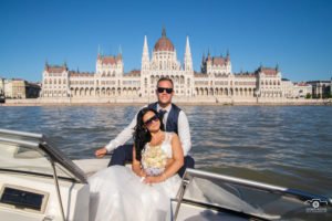 Romantikus esküvői fotózás Budapesten, a Duna vizén. A boldog pár stílusos napszemüvegben élvezi a hajózást, háttérben a magyar Parlament impozáns épületével, amely lenyűgöző díszletet biztosít a különleges alkalomhoz.
