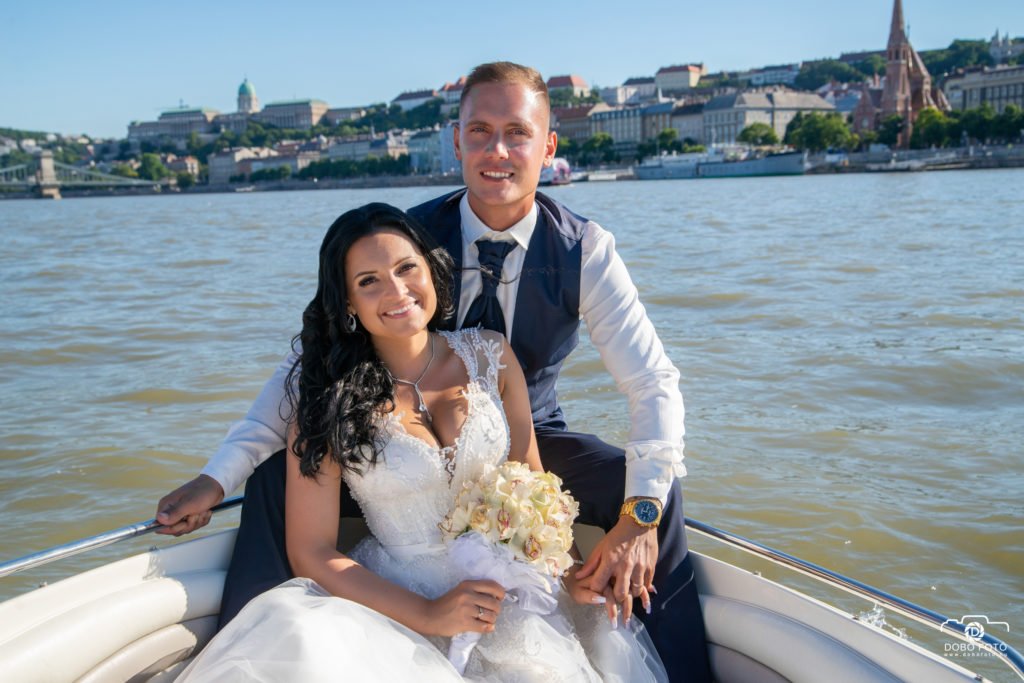 Esküvői fotózás Budapesten, a Duna vizén hajókázva. A boldog pár romantikus és természetes pózban mosolyog a kamera felé, háttérben a budai vár látványával, amely tökéletesen kiegészíti az esküvő különleges hangulatát.