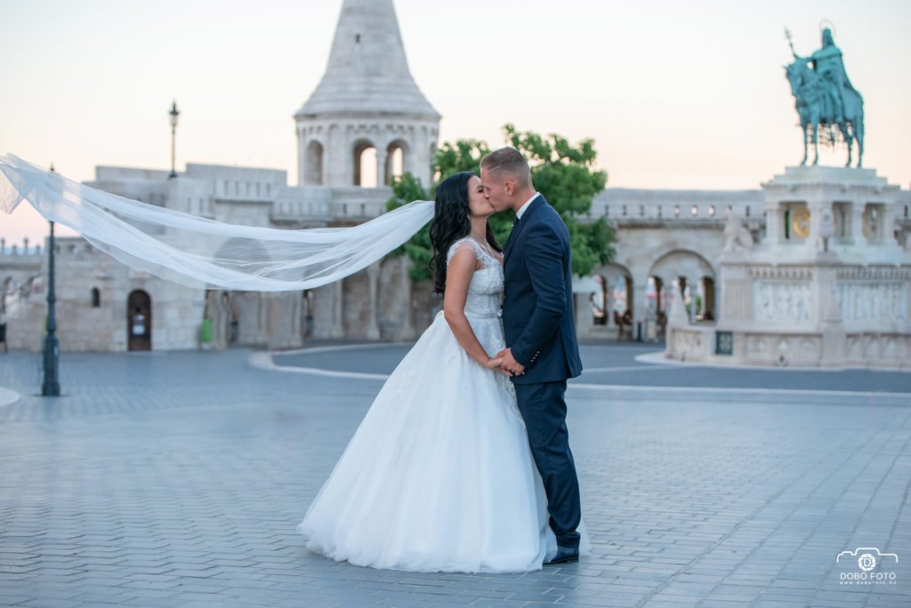 Esküvői csók a budapesti Halászbástya előtti téren, ahol a menyasszony fátyla elegánsan lebeg a szélben. A Hajdúböszörményből érkezett fotós különleges pillanatot örökített meg a pár kreatív esküvői fotózásán, kihasználva a lenyűgöző történelmi helyszín varázsát.