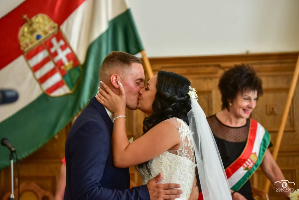A polgári esküvő megható pillanata, amikor az ifjú pár megosztja első csókját házaspárként. A menyasszony gyönyörű csipkés fehér ruhában, fátyollal, a vőlegény elegáns kék öltönyben látható. A háttérben a magyar nemzeti zászló és az anyakönyvvezető mosolyogva figyeli az eseményt, amely Hajdúböszörmény ünnepi hangulatát idézi. Az esküvői fotós profi módon örökítette meg ezt a romantikus és emlékezetes pillanatot.