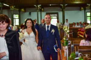 Az ifjú pár kéz a kézben lépdel az esküvői szertartás során a templomban, sugárzó mosollyal az arcukon. A menyasszony elegáns, csipkézett esküvői ruhában, a vőlegény klasszikus kék öltönyben tündököl. A háttérben az ünneplő násznép látható, az esküvői fotós tökéletesen örökítette meg a pillanatot, amely a nap romantikus és meghitt hangulatát idézi.