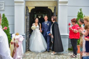 A boldog pár kilép a templom kapuján, kéz a kézben, mosollyal az arcukon, miközben a vendégek virágszirmokkal várják őket. Az esküvői fotós ezt a meghitt pillanatot örökítette meg, kiemelve a ceremónia ünnepélyes és romantikus hangulatát.