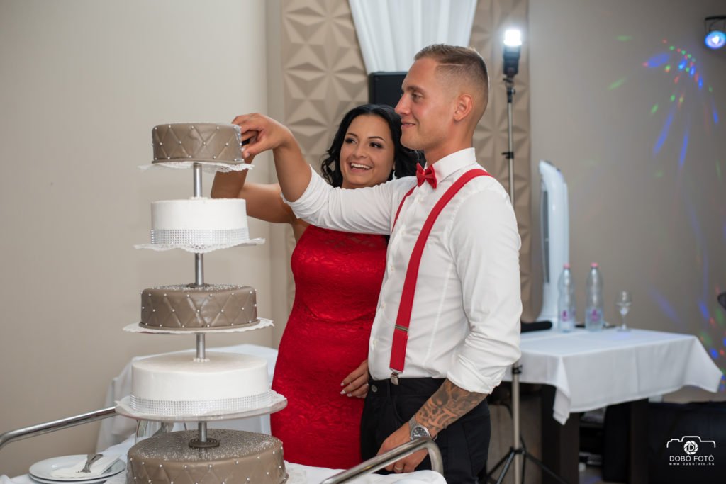 Esküvő fotós - Hajdúböszörmény. Egy pár éppen az esküvői torta felszeletelésére készül egy elegáns eseményen Hajdúböszörményben. A többemeletes torta különleges díszítéssel látható, miközben a pár mosolyogva élvezi a pillanatot.