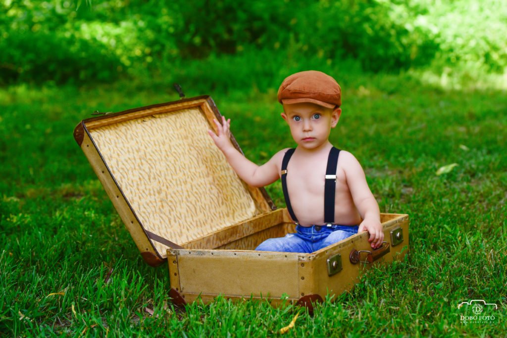 Kisgyermek vintage bőröndben ülve a zöld természet közepén, kalapban és nadrágtartóval. Kreatív kültéri gyermekfotózás Debrecenben, nosztalgikus hangulatú beállítással.