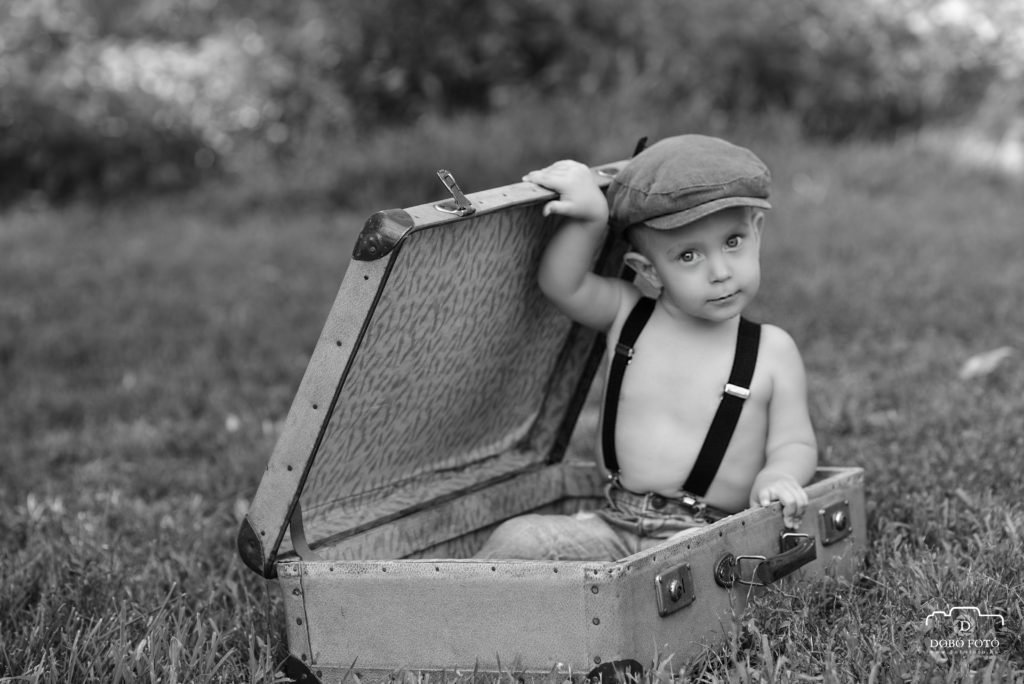 Kisgyermek fekete-fehér fotó vintage bőrönddel a természetben, kalapban és nadrágtartóval. Kreatív kültéri gyermekfotózás Debrecenben, klasszikus és időtálló hangulatban.