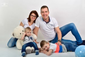 Vidám családi műtermi fotó: szülők két kisfiúval és nagy fehér plüssmackóval, egységes fehér-farmer öltözetben, kék léggömbbel, fehér háttér előtt. DOBO FOTO családi fotózás.