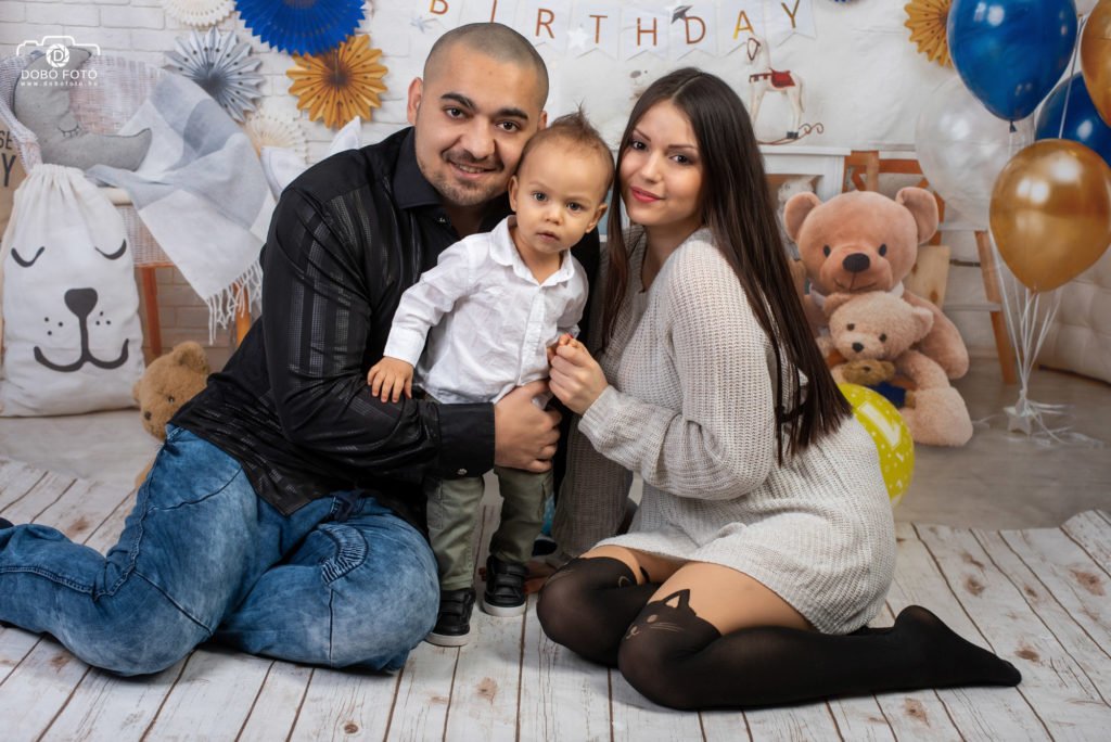 Családi fotó születésnapi díszlettel: szülők kisfiukkal dekorált háttér előtt, lufikkal és plüssmackókkal, fehér fa padlón ülve. DOBO FOTO stúdiófotózás.