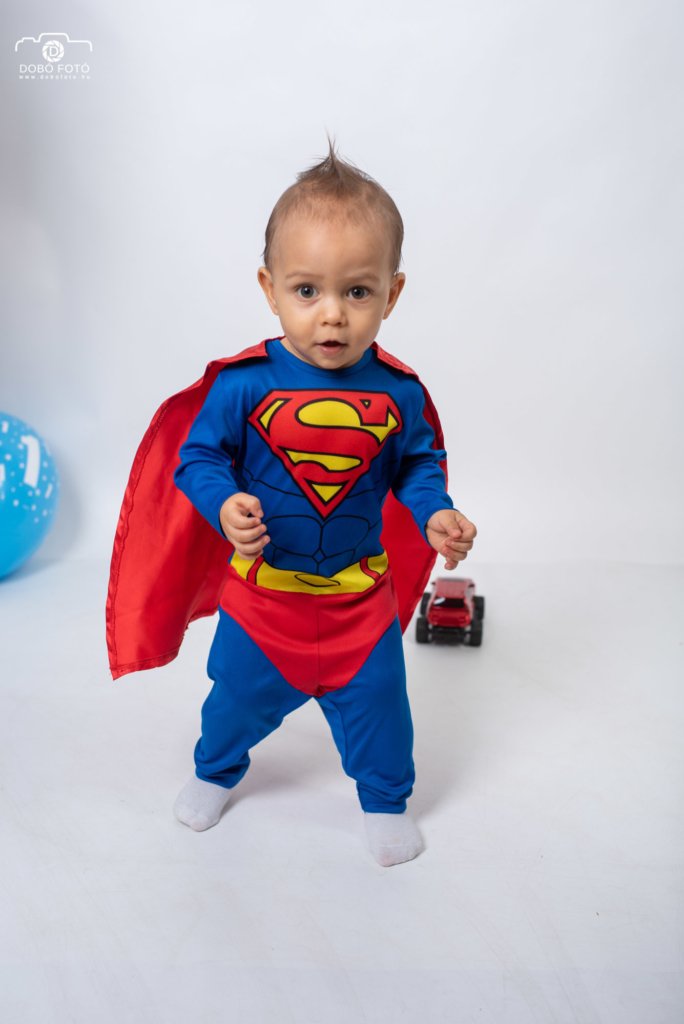 Kisfiú Superman jelmezben, piros köpenyben, kék játékautóval a háttérben, fehér műtermi környezetben. Dobó Fotó gyermek fotózás.