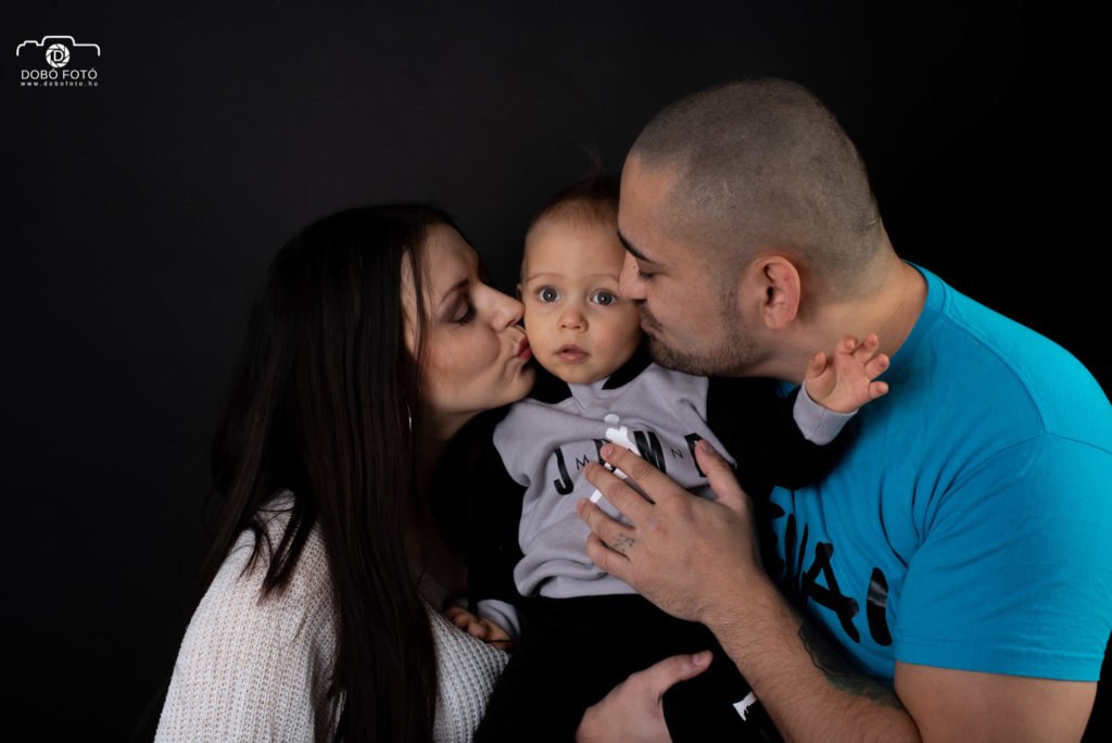 Intim családi portré fekete háttér előtt: szülők puszit adnak kisbabájuk arcára, apa türkiz pólóban, anya fehér kötött pulóverben. DOBO FOTO műtermi fotózás.