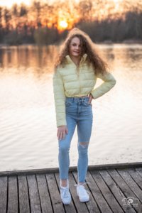 Szabadtéri portré. Fiatal lány világos sárga dzsekiben és koptatott farmerben áll egy tóparti stégen, háttérben a naplemente aranyló sugarai világítják meg a tájat.