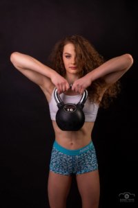 Dinamikus fitnesz fotó: fiatal sportoló fekete háttér előtt, fehér topban és kék mintás rövidnadrágban, kettlebell súlyzóval edző pózban. DOBO FOTO debreceni portréfotózás.
