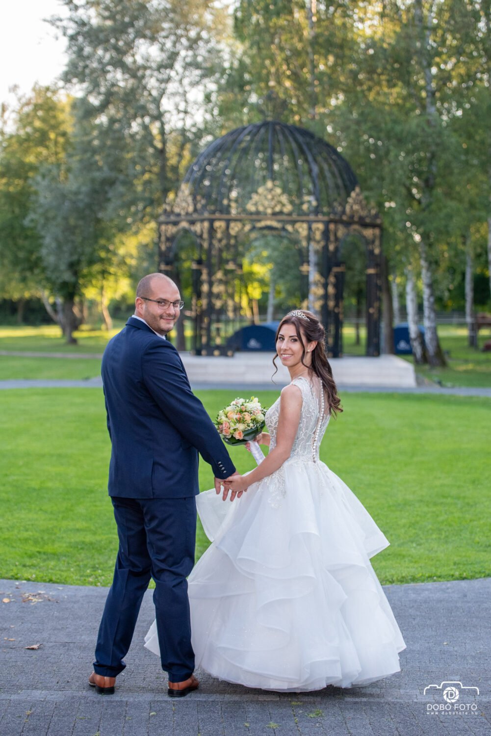 Esküvői fotózás a Park Hill varázslatos kertjében, ahol a boldog pár kézenfogva sétál, háttérben az elegáns pavilonnal és a természet zöldellő szépségével.