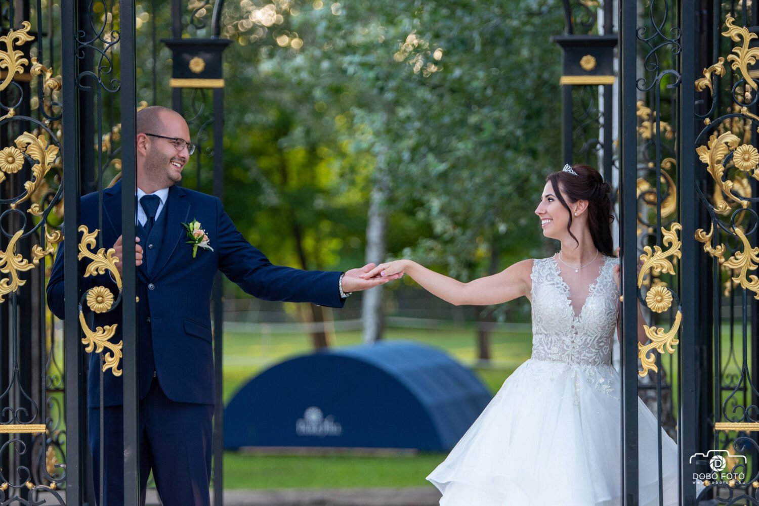 Esküvői fotó, ahol a boldog pár kézfogása egy elegáns kovácsoltvas kapu két oldalán történik, romantikus pillanat a természetben.