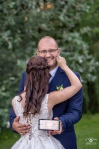 Esküvői portré egy vidám pillanattal, a vőlegény kezében egy 'HELP!' üzenettel, miközben a menyasszony átöleli őt romantikus környezetben.