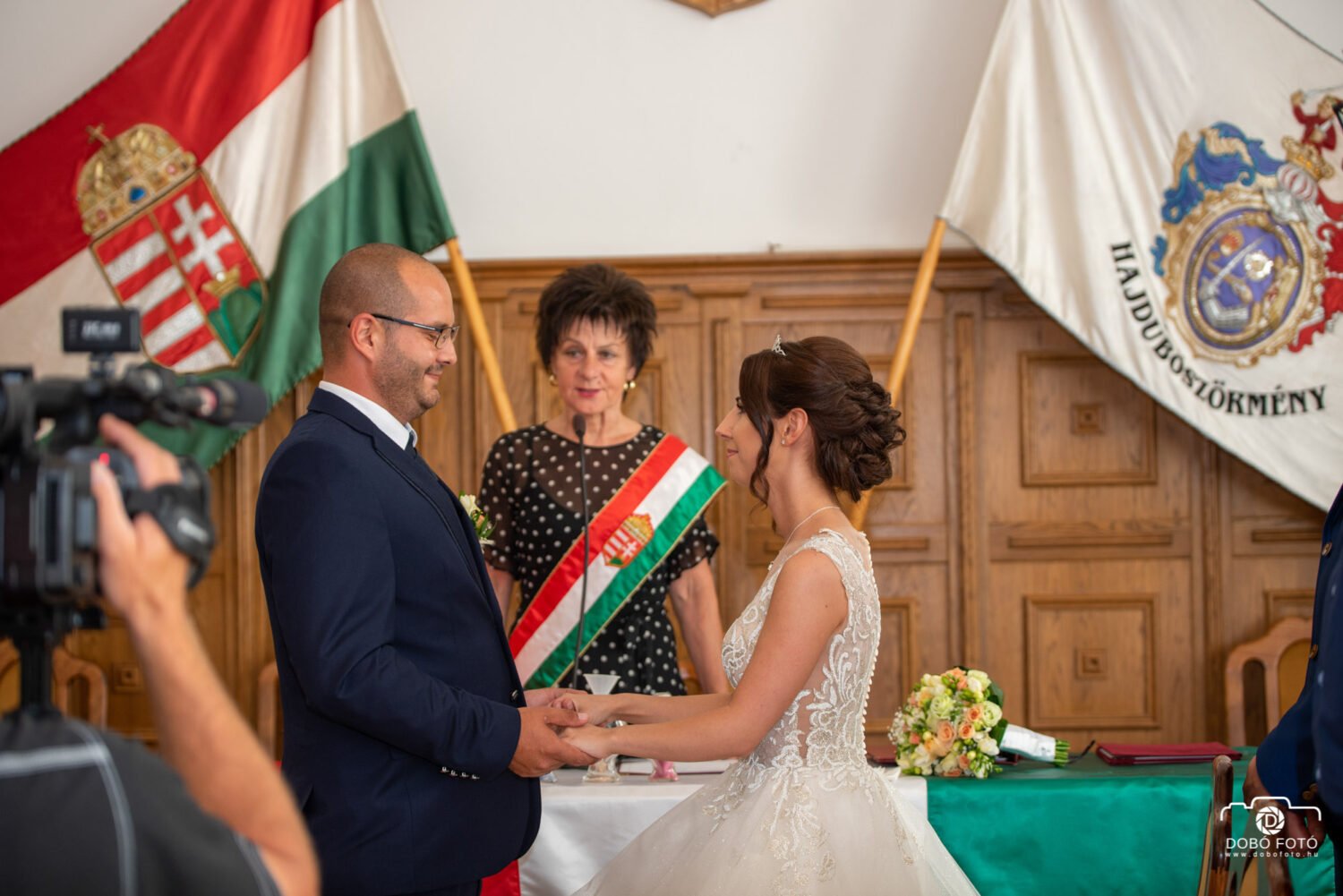 Esküvő fotózás - Debrecen. A menyasszony és a vőlegény a polgári szertartás alatt fogadalmat tesz Hajdúböszörményben, Magyarország zászlói és címer előtt.