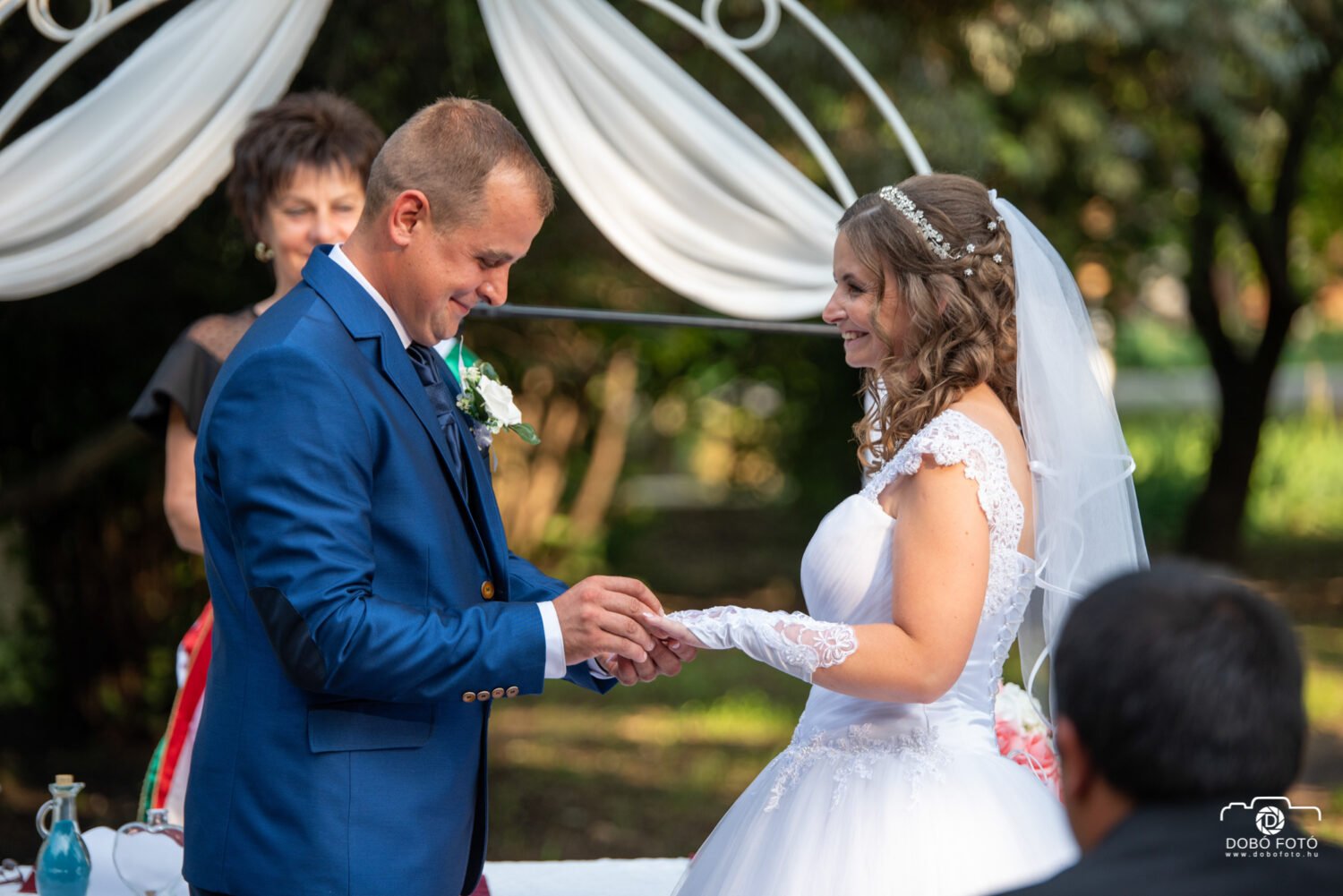 Egy menyasszony és vőlegény mosolyogva húzzák fel egymás ujjára a gyűrűt egy szabadtéri esküvői szertartáson Hajdúviden. A menyasszony elegáns fehér csipkeruhát és fátylat visel, míg a vőlegény kék öltönyben áll mellette. A háttérben természetes környezet és esküvői dekoráció látható, amely meghitt hangulatot teremt.