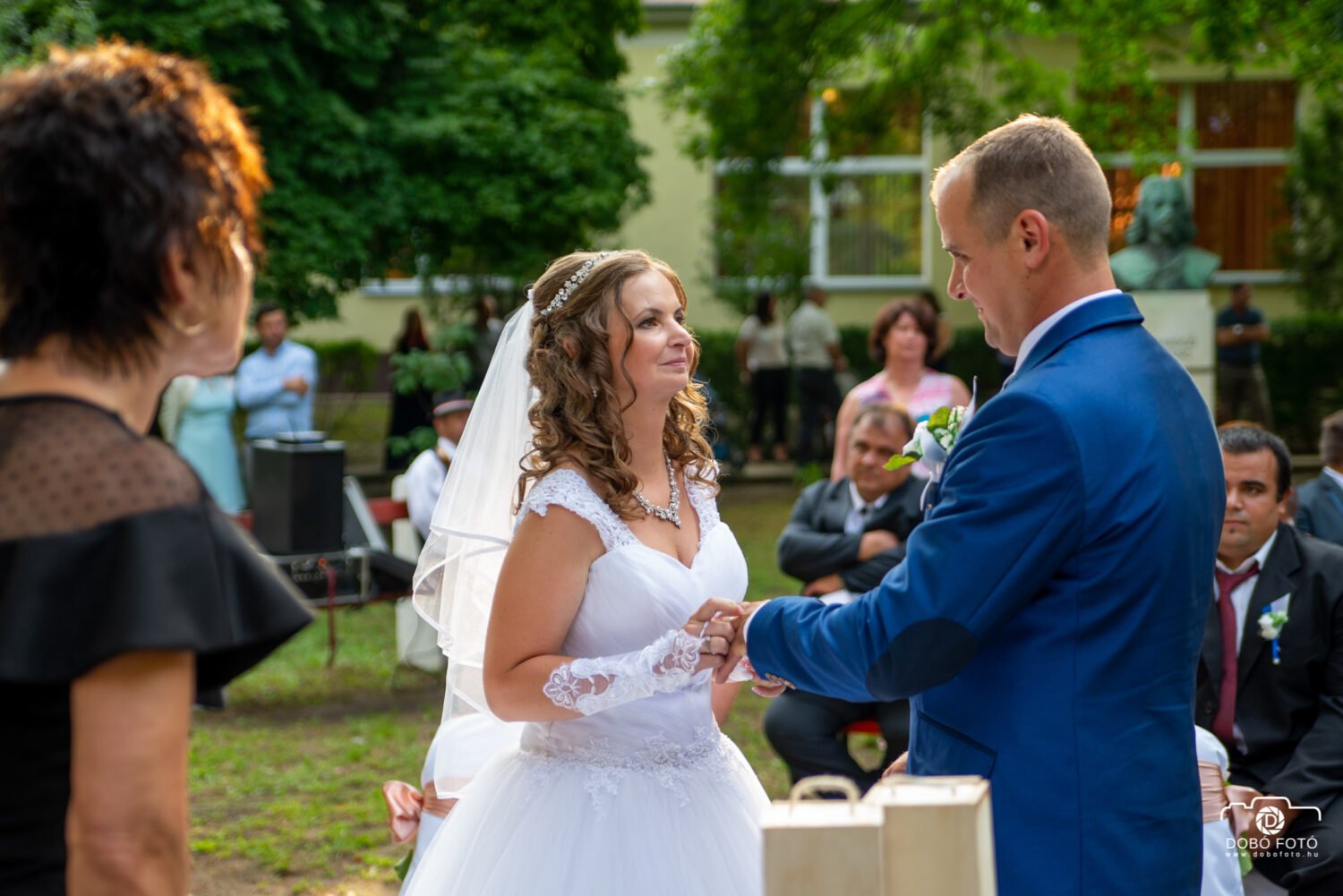 Egy menyasszony és vőlegény tartja egymás kezét a fogadalomtétel közben egy szabadtéri esküvői szertartáson Hajdúviden. A menyasszony elegáns fehér csipkeruhát visel fátyollal és gyöngyös hajdísszel, míg a vőlegény kék öltönyben áll mellette. A háttérben vendégek és természetes környezet teremtenek meghitt hangulatot az eseményhez.