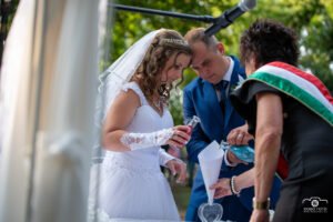 Esküvői fotós - Debrecenben. Egy menyasszony és vőlegény homokceremóniát végez az esküvői szertartás során, színes homokot öntve egy közös edénybe, amely a házasság egységét jelképezi. A menyasszony elegáns fehér csipkeruhát, a vőlegény pedig kék öltönyt visel. Az anyakönyvvezető nemzeti szalagot visel, amely a ceremónia hivatalos jellegét emeli ki. A háttérben zöld természet ad kellemes hangulatot a szabadtéri eseménynek.