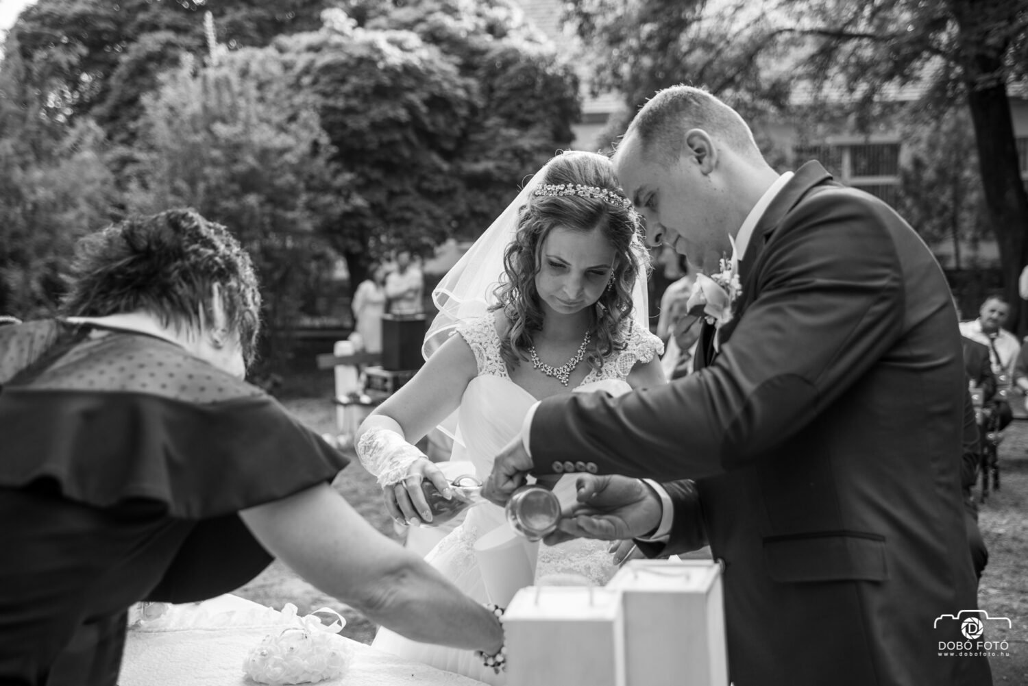 Egy menyasszony és vőlegény közösen önti a színes homokot egy edénybe a homokceremónia részeként, amely a házasság egységét szimbolizálja. A fekete-fehér fotó kiemeli a menyasszony elegáns csipkeruháját és fátylát, valamint a vőlegény klasszikus öltönyét. A ceremónia meghitt hangulatát a háttérben lévő természet és ünneplő vendégek teszik teljessé.
