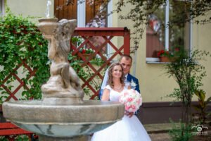 Egy mosolygó menyasszony és vőlegény áll a háttérben egy szökőkúttal, amely romantikus hangulatot kölcsönöz az esküvői fotónak. A menyasszony elegáns fehér csipkeruhát visel rózsaszín és fehér virágokból álló csokorral a kezében, míg a vőlegény kék öltönyben öleli át. A vörös rácsos háttér és zöld növények harmonikusan keretezik a jelenetet.