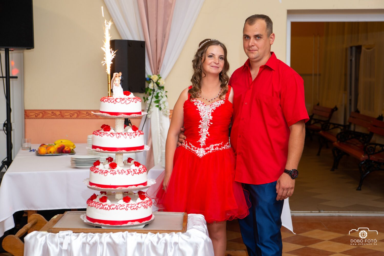 Menyecsketánc pillanata egy esküvőn, ahol az ifjú pár az ünnepi, piros-fehér esküvői torta mellett áll. A menyasszony piros menyecskeruhában, a vőlegény piros ingben mosolyog. Esküvői fotózás, kreatív esküvői képek és különleges pillanatok megörökítése professzionális esküvői fotós által.