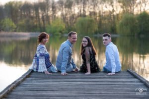 Egy négytagú család ül a Vekeri-tó partján egy fából készült stégen, miközben hátrafordulnak a kamerába. A szabadtéri fotózás során a természet nyugodt szépsége és a tó tükröződése harmonikus hátteret biztosít, kiemelve a család összhangját és a pillanat meghittségét.