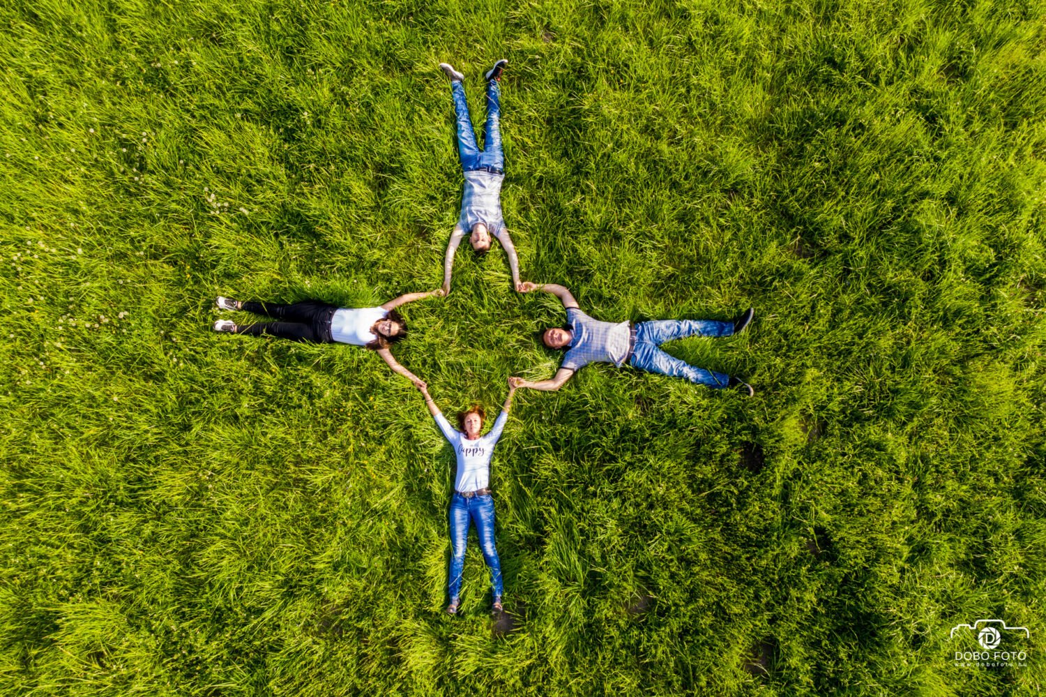 Szbadtéri családi fotózás.Egy négytagú család fekszik a zöld fűben, csillag alakzatot formálva, miközben kezeiket összekulcsolják. A felvétel madártávlatból készült, a természet zöldje harmóniát sugároz a családi összhanggal.