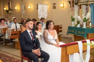 Esküvői szertartás az újfehértói katolikus templomban, ifjú pár első sorban, fehér gladióluszok az oltárnál, bensőséges pillanat.