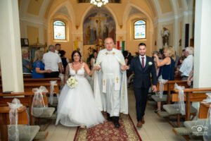 Katolikus templomi szertartás Újfehértón, ifjú pár és plébános a főhajóban, díszes oltár és színes üvegablakok, esküvői fotózás művészi megörökítése.