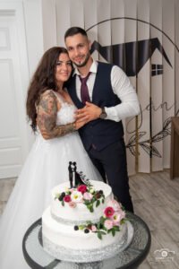 Ifjú pár az esküvői tortájuk mellett, fehér kétemeletes torta rózsaszín virágdíszítéssel, modern stúdiószettek előtt, menyasszony tetovált karral, vőlegény mellényben.