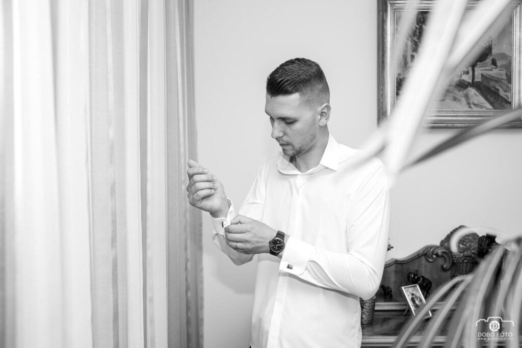 A vőlegény gondosan begombolja az ingét, miközben készülődik a nagy napra. Elegancia és részletekre való odafigyelés az esküvői pillanatokban.