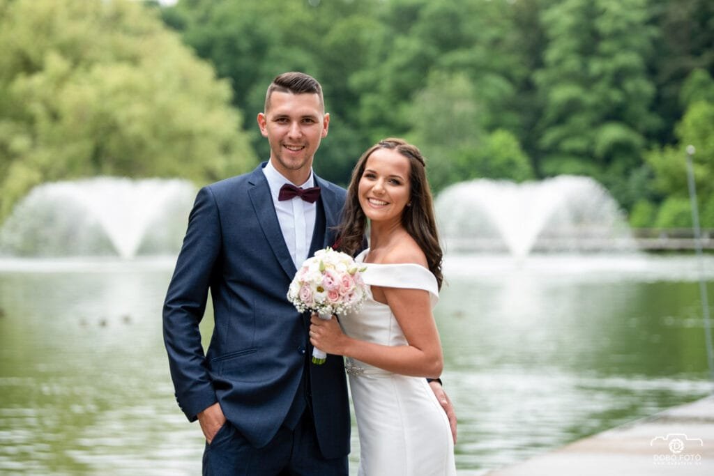 A boldog ifjú pár esküvői fotója a debreceni Nagyerdő festői szökőkútjának háttérében. Elegáns öltözetben, virágcsokorral a kezükben, harmonikus és romantikus pillanat.
