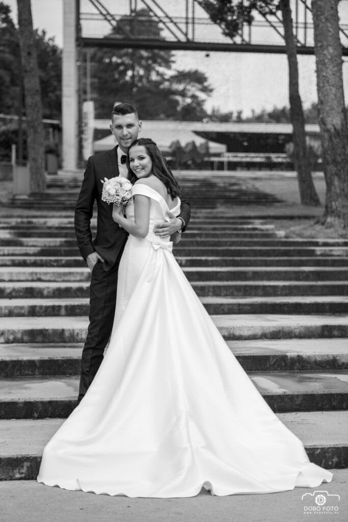 Menyasszony és vőlegény szorosan egymás mellett, mosolyogva állnak a Debreceni Nagyerdőben, háttérben lépcsősorral. Elegáns és meghitt esküvői fotó klasszikus fekete-fehér stílusban.