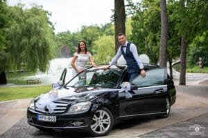 Menyasszony és vőlegény díszített autóval a Debreceni Nagyerdő idilli környezetében, háttérben egy szökőkúttal. Stílusos és meghitt esküvői pillanat.