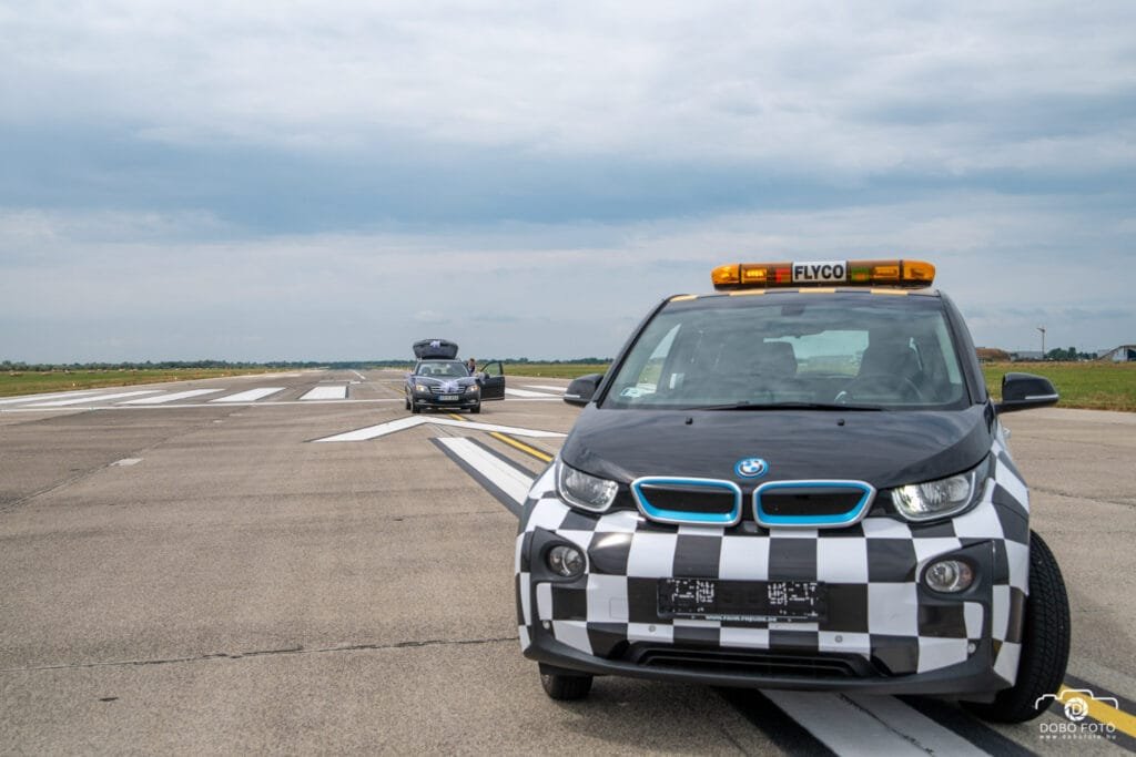BMW FLYCO jármű a repülőtér kifutópályáján, háttérben egy esküvői dekorációval ellátott autóval. Egyedi esküvői fotózás különleges környezetben és kísérettel.