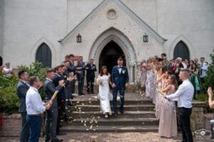 Menyasszony és vőlegény kéz a kézben lépnek ki a templomból, miközben konfetti záporral ünnepli őket a násznép. Meghitt és elegáns esküvői pillanat.