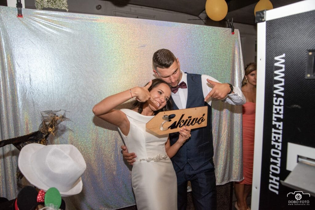 Selfiebox bérlés - Debrecen. Menyasszony és vőlegény a selfiebox előtt, kezükben "Esküvő" feliratú dekorációval. Szórakoztató esküvői fotózás kreatív kiegészítőkkel és háttérrel.
