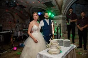 Esküvő fotózás - Dobófotó. A menyasszony és a vőlegény az esküvői torta mellett állnak, amelyet csillagszóró díszít. A háttérben a Hotel Óbester hangulatos boltíves terme látható, színes fényekkel és zenei eszközökkel.