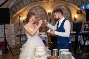 A menyasszony és a vőlegény közösen kínálják egymást az esküvői tortából a debreceni Hotel Óbester boltíves termében. A háttérben hangszerek és fények teszik teljessé az ünnepi hangulatot.