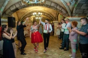Esküvő fotózás - Debrecen. Vidám esküvői jelenet a debreceni Hotel Óbester egyik boltíves termében. A menyasszony piros szoknyában és hímzett felsőben, a vőlegény piros nadrágtartóban és fehér ingben táncol a násznép tapsa és nevetése közepette.