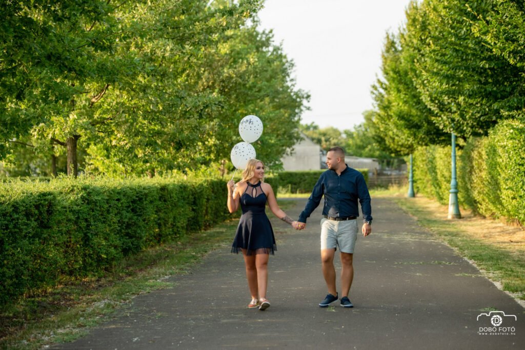 Jegyespár sétája naplemente előtt, kék ruhás pár lufival, zöld fás park háttérrel.