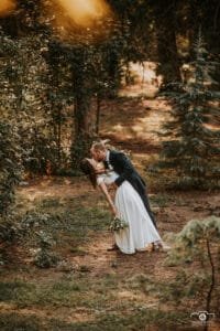 Esküvői kreatív fotózás -Dobó Fotó. Romantikus esküvői fotó, ahol a vőlegény hátradönti a menyasszonyt, és szenvedélyesen megcsókolja, miközben a menyasszony egy virágcsokrot tart. Az erdő természetes fényével és zöldellő fáival a háttérben varázslatos hangulatot áraszt.